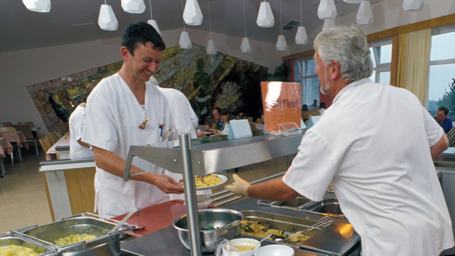 Zwei Männer in weißer Krankenhaus-Kleidung an einer Essensausgabe in einer Kantine