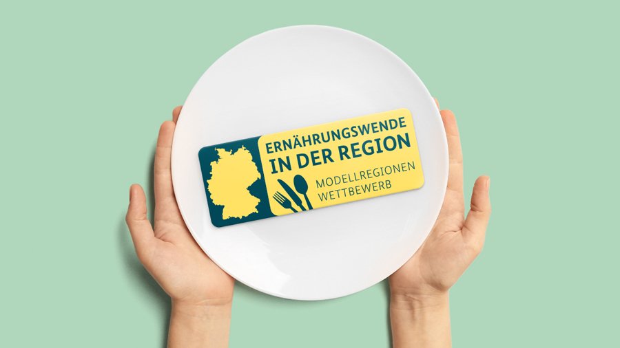Zwei Hände halten einen hellgrünen Teller mit dem Logo des Modellregionenwettbewerbs; Text: Ernährungswende in der Region - Modellregionenwettbewerb 